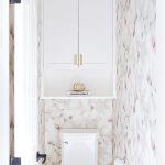 Chic water closet is clad in pink and gray floral wallpaper lined with  a cabinet and shelf hovering over a toilet illuminated by a Soleil Small  Pendant.
